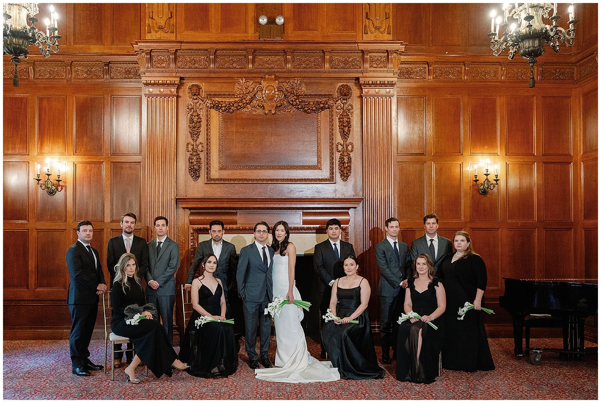 The full wedding party poses together in one of the Harmonie Club's grand Manhattan rooms. Groomsmen in dark suits and bridesmaids in black dresses flank the bride and groom, with the club's signature rich wood paneling and wall sconces behind them.