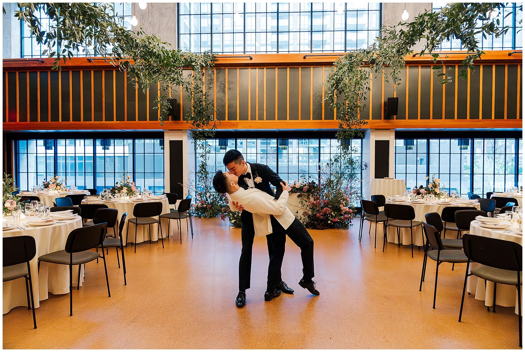 One groom in a white tuxedo jacket dips his partner in their reception space at the Ace Hotel Brooklyn, New York. The lofty industrial space is filled with sweeping greenery and wedding florals hanging overhead, creating a dreamy NYC wedding reception backdrop.