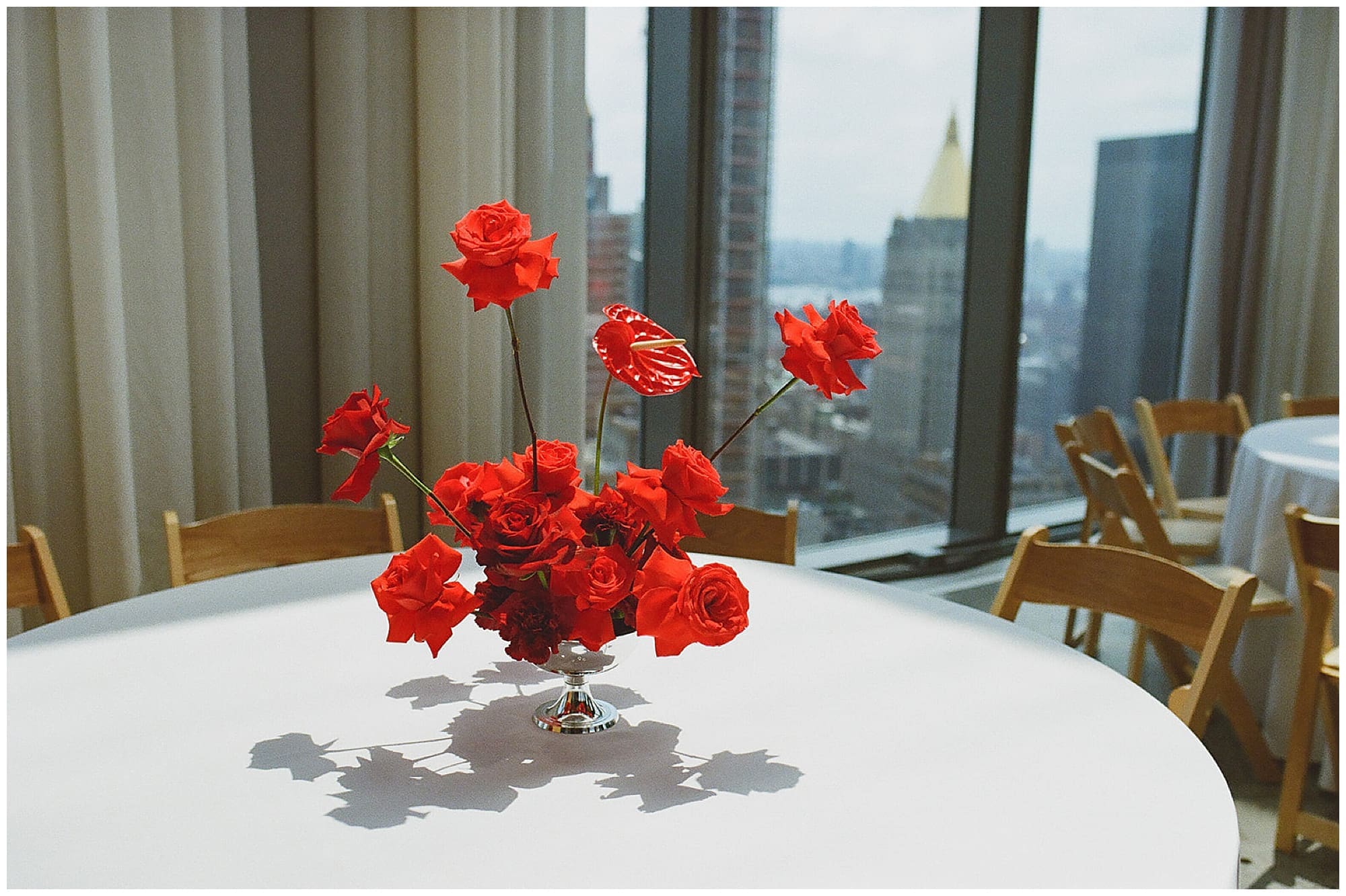 A sculptural red carnation and rose floral centerpiece sits on a white table at Virgin Hotels NYC, with the Manhattan skyline visible through the floor-to-ceiling windows behind it, styled by a New York City wedding florist.