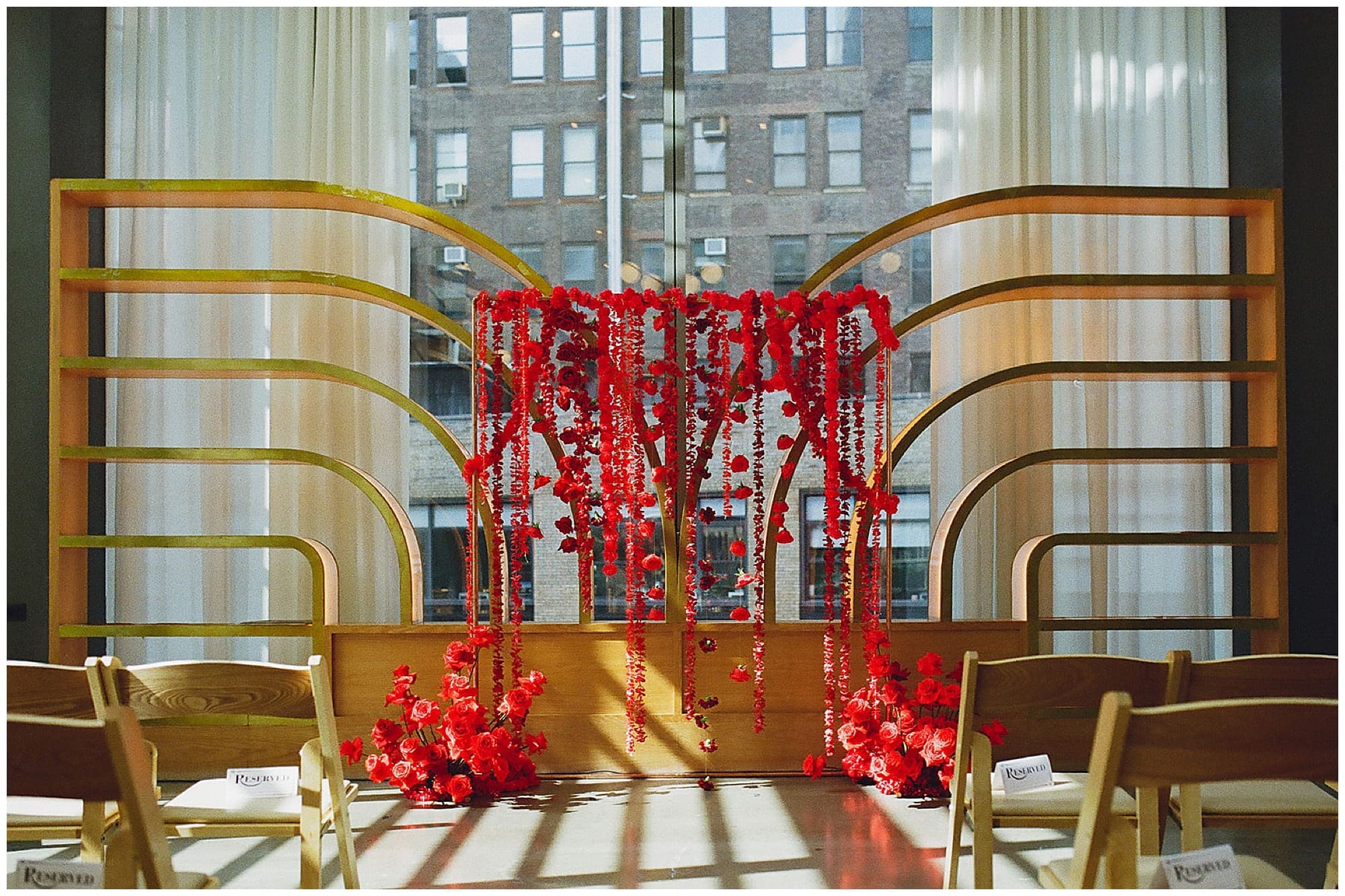 A wide shot of the Hindu ceremony space at Virgin Hotels NYC in Manhattan. A gold arch mandap adorned with cascading red floral strings and beaded garlands is flanked by large windows with sweeping New York City views, with guests seated on either side.