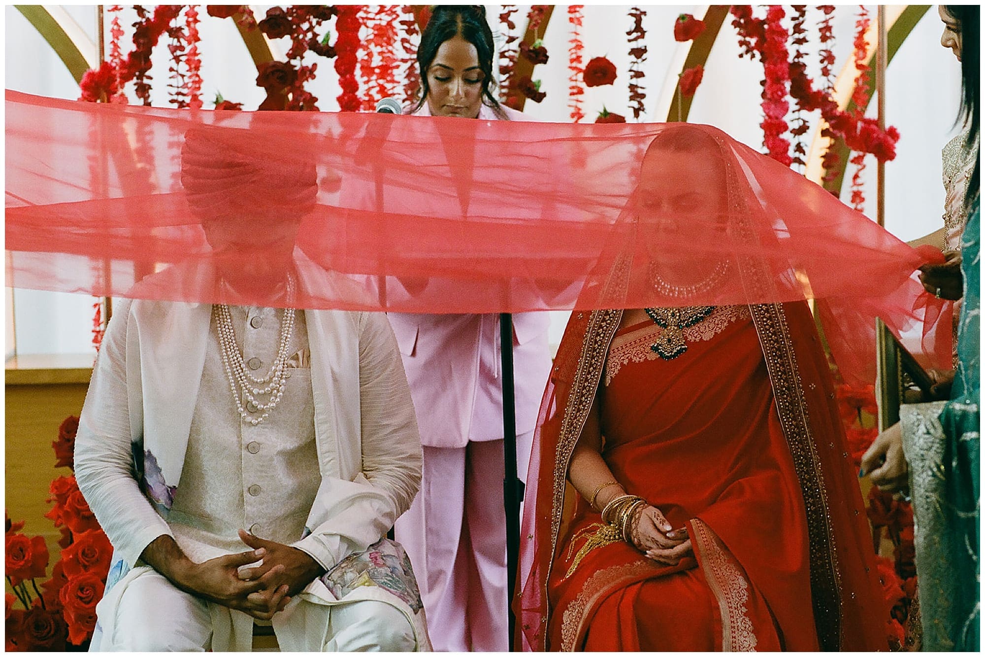 The groom sits during a ceremony ritual beneath a sweeping red fabric canopy at Virgin Hotels NYC, dressed in a white sherwani and surrounded by cascading red wedding florals. A tender and sacred moment from this Manhattan Hindu wedding.