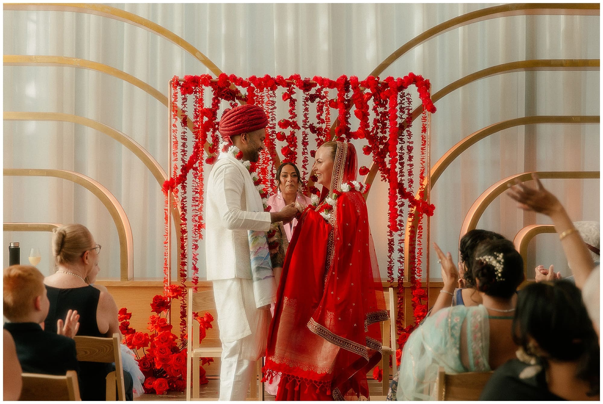 The ceremony space at Virgin Hotels NYC in Manhattan is set for a Hindu wedding, with a gold arch mandap decorated with cascading red floral garlands and beads, surrounded by seated guests in a bright, modern New York City event space.