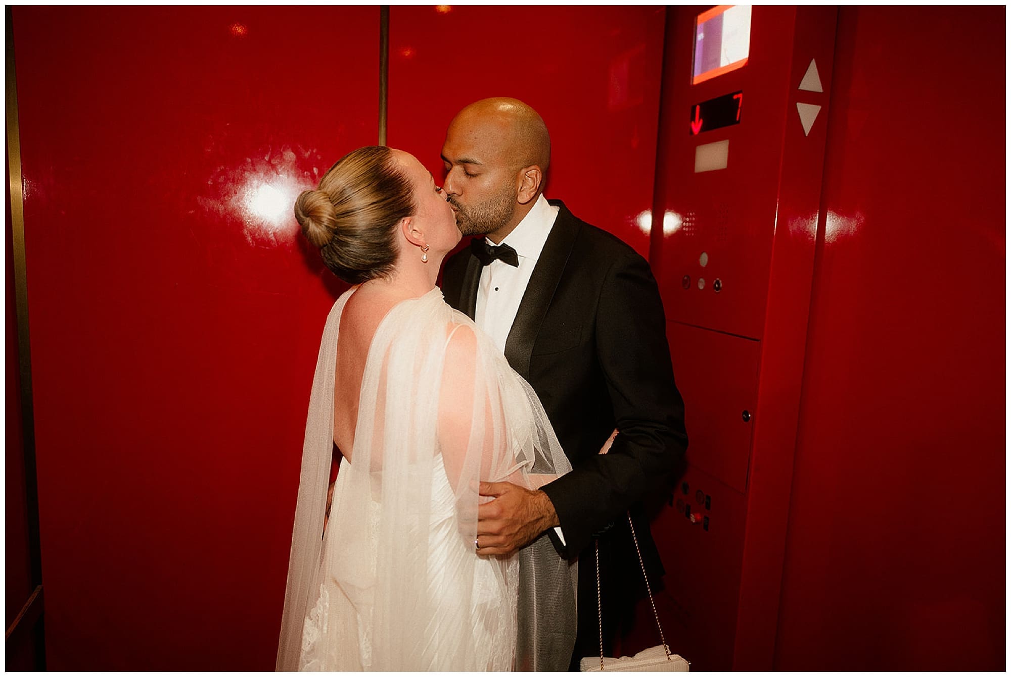 The couple shares a kiss in front of a dramatic red lacquered wall at Virgin Hotels NYC, creating a bold and romantic portrait on their Manhattan wedding day.