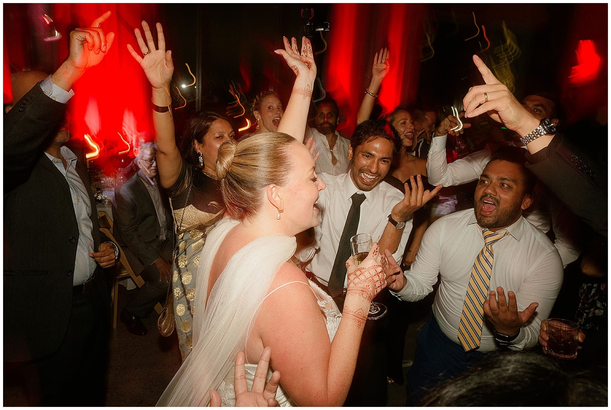 The bride throws her arms up in celebration on the dance floor at Virgin Hotels NYC, surrounded by cheering guests beneath green and red reception lighting. Pure joy from this New York City wedding night.