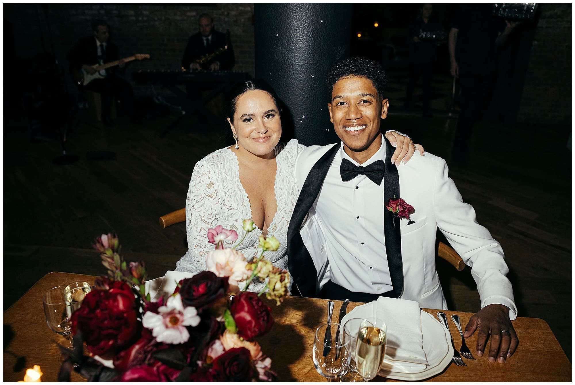 The bride and groom sit together at the reception table at the Wythe Hotel Brooklyn, both dressed in white, with a lush arrangement of deep red and burgundy wedding florals between them, smiling at the camera on their NYC wedding day.