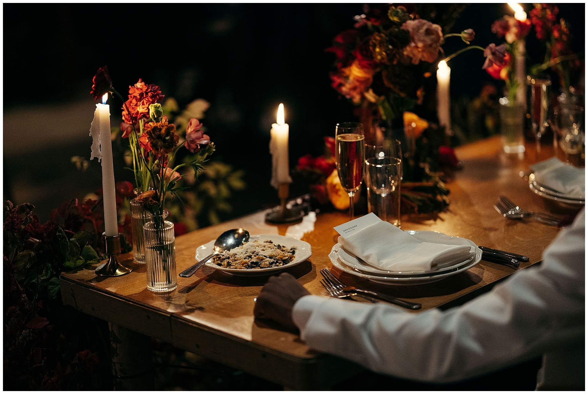 The long reception table at the Wythe Hotel in Brooklyn, New York, is styled with dramatic floral centerpieces in deep red and jewel tones, tall taper candles, and rich textiles, creating a moody and elegant New York City wedding atmosphere.