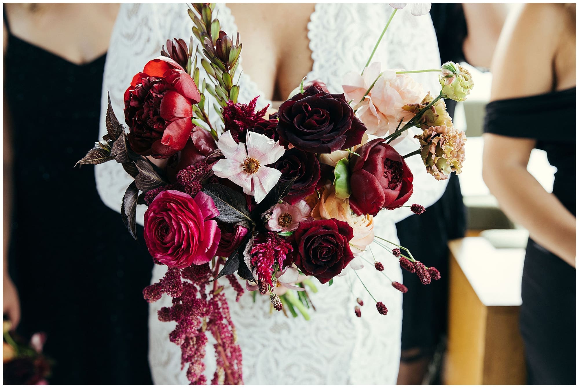 A close-up detail of the bride's hands holding a dramatic cascading bridal bouquet overflowing with deep red roses, burgundy blooms, and blush and peach florals, created by a NYC wedding florist.