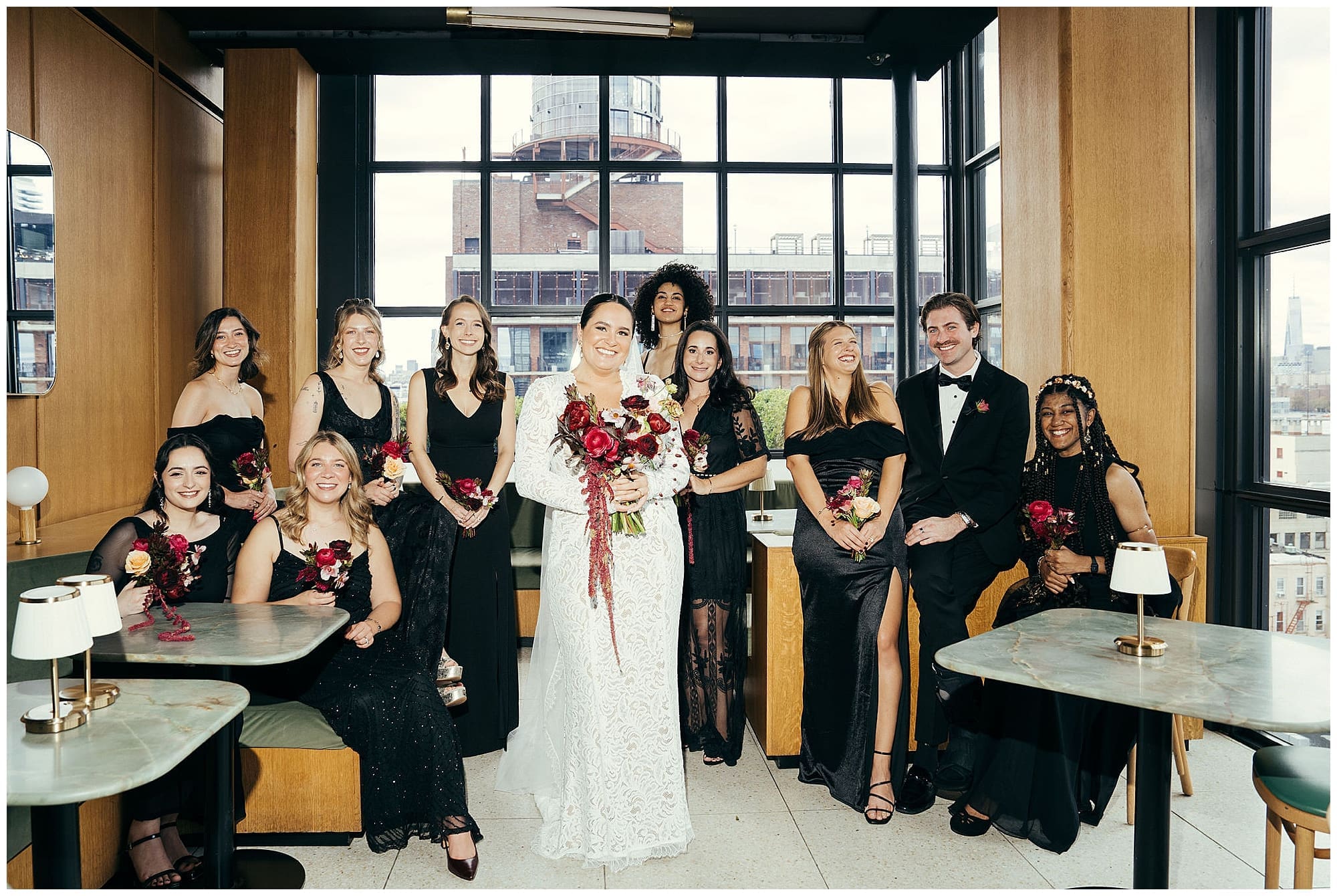 The wedding party gathers indoors at the Wythe Hotel Brooklyn for a group portrait, bridesmaids in black dresses flanking the couple in front of large industrial windows on this New York City wedding day.