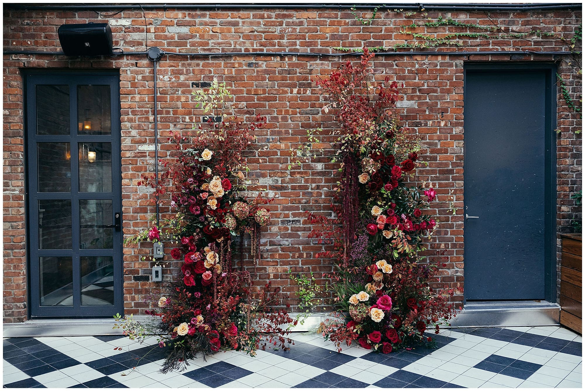 A sweeping floral installation of deep red, burgundy, blush, and peach wedding florals cascades against the exposed brick wall at the Wythe Hotel in Brooklyn, New York, created by the couple's New York City wedding florist.