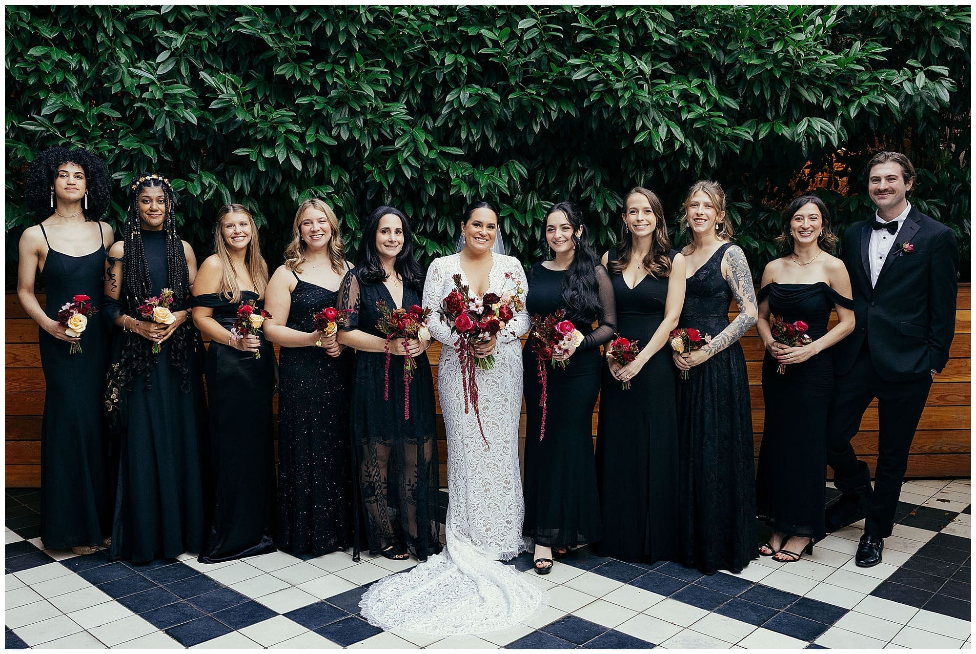 The bridesmaids in black gowns and the couple pose together on the iconic black and white checkered floor at the Wythe Hotel in Brooklyn, New York, surrounded by lush greenery.