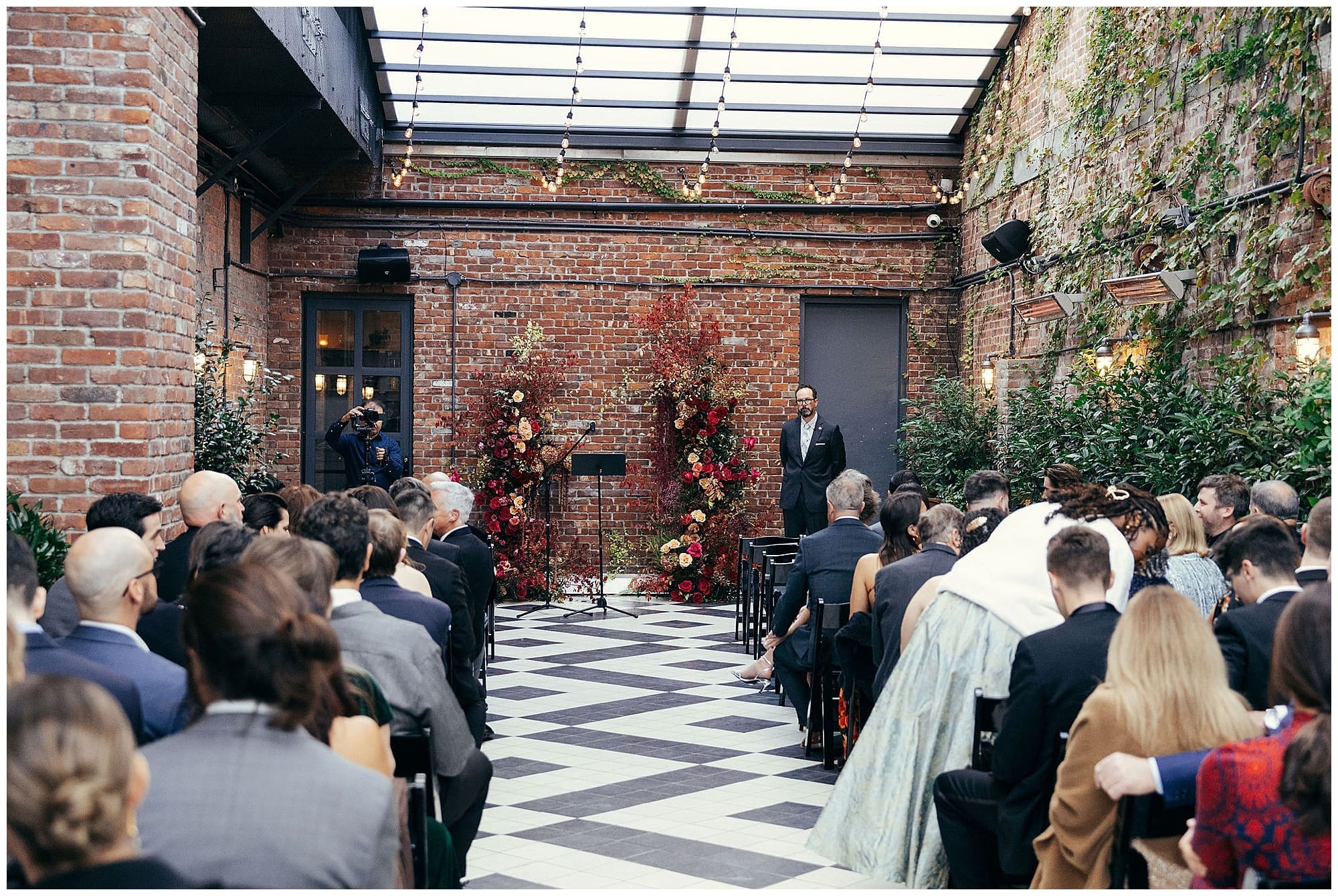 A wide shot of the ceremony space at the Wythe Hotel in Brooklyn, NYC, with guests seated on the black and white checkered floor and a lush floral arch of deep red and burgundy wedding florals framing the couple at the altar.