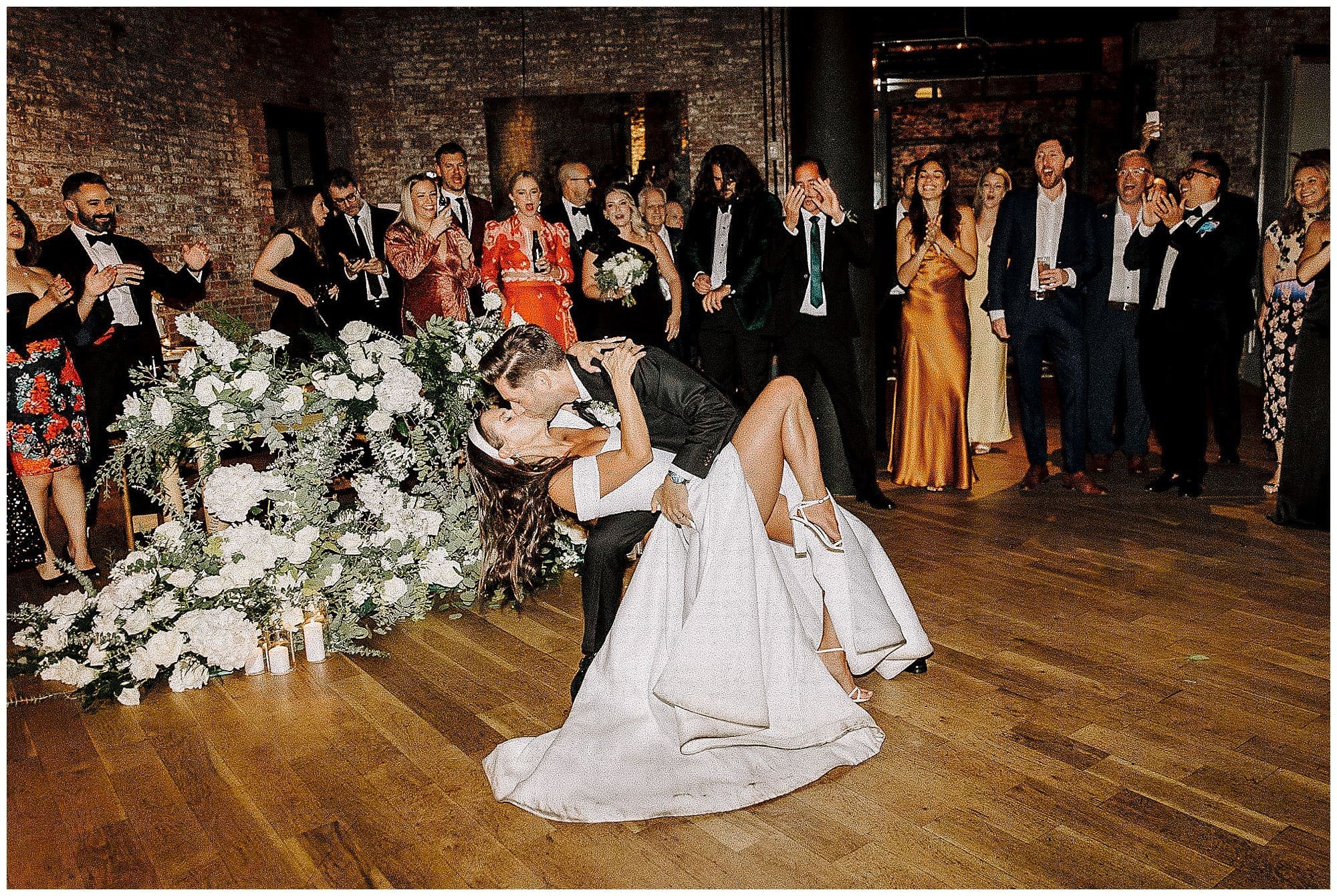 The groom dips the bride dramatically on the dance floor during their first dance at the Wythe Hotel in Brooklyn, NYC, surrounded by cheering guests. The bride's white gown fans out as she arches backward, with lush wedding florals visible throughout the reception space.