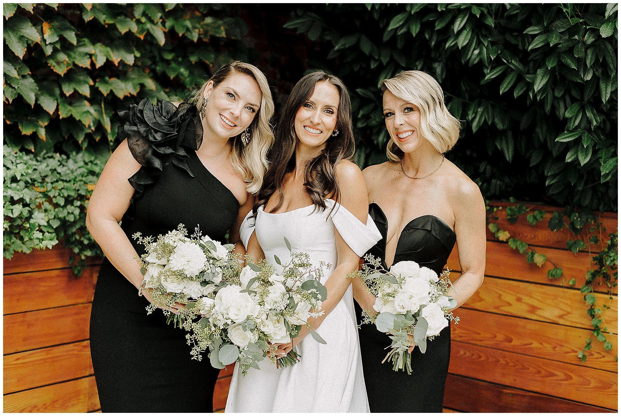 The bride stands with two bridesmaids in black gowns in front of a lush ivy-covered wall at the Wythe Hotel in Brooklyn, NYC. All three hold white and green wedding florals and smile toward the camera.