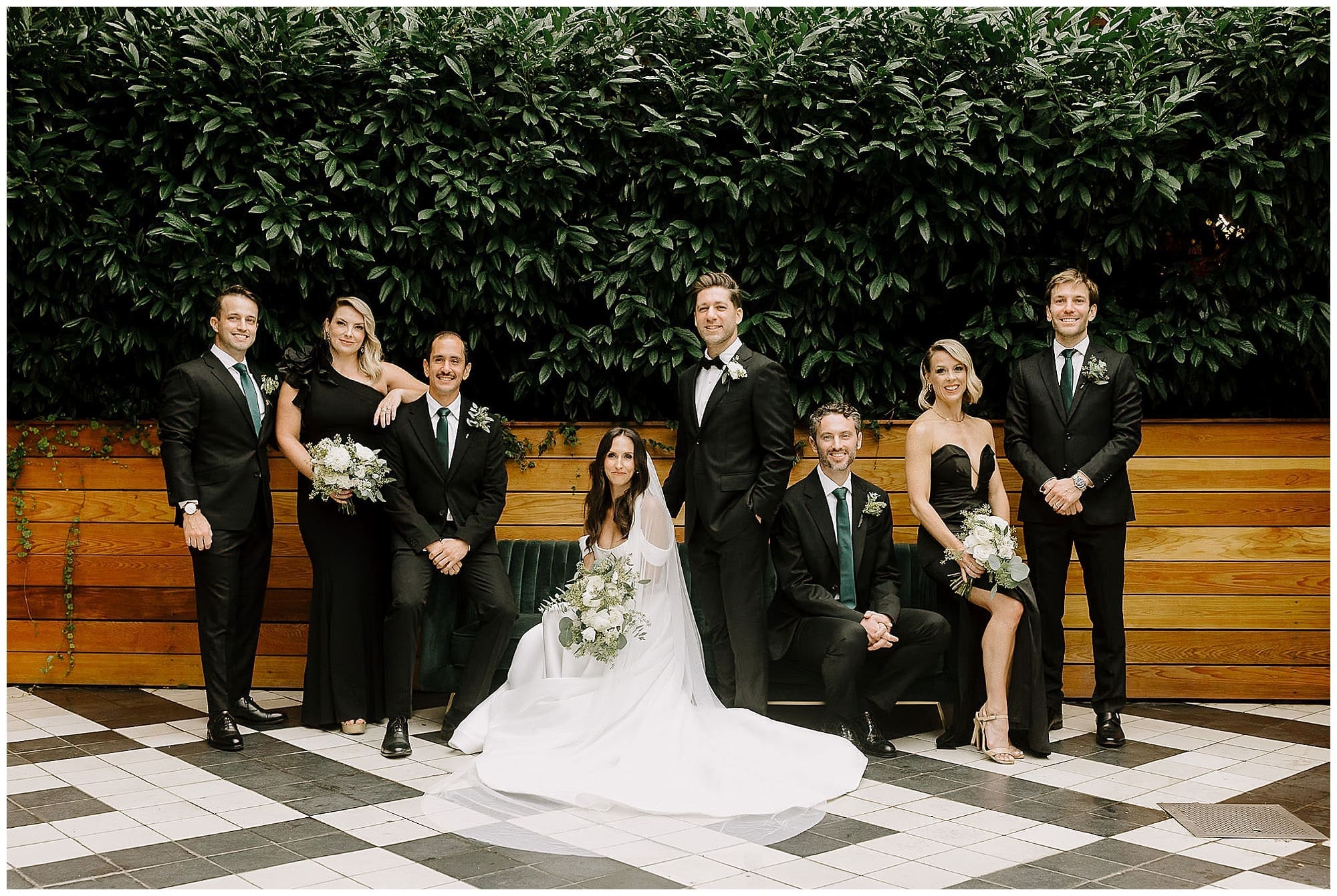 The wedding party poses together on the black and white checkered floor at the Wythe Hotel, Brooklyn, New York. The bride sits in the center in her white gown, surrounded by the groom and wedding party in black attire, with greenery and wedding florals in the background.