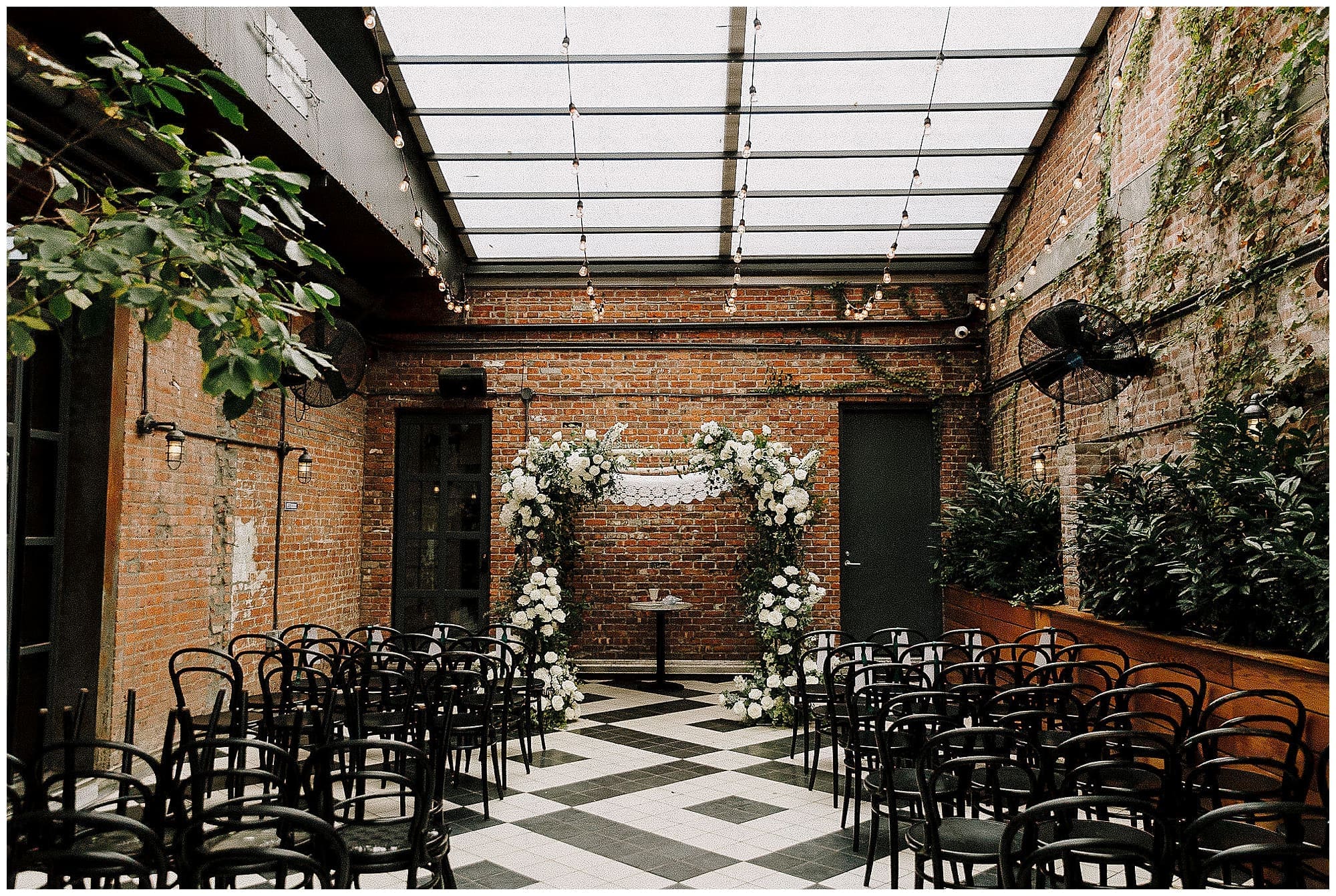 An empty ceremony space at the Wythe Hotel in Brooklyn, NYC, with a black and white checkered floor. Black bistro chairs are arranged in rows facing a stunning floral arch created by a New York wedding florist, adorned with white flowers and greenery beneath a glass ceiling with exposed brick walls.