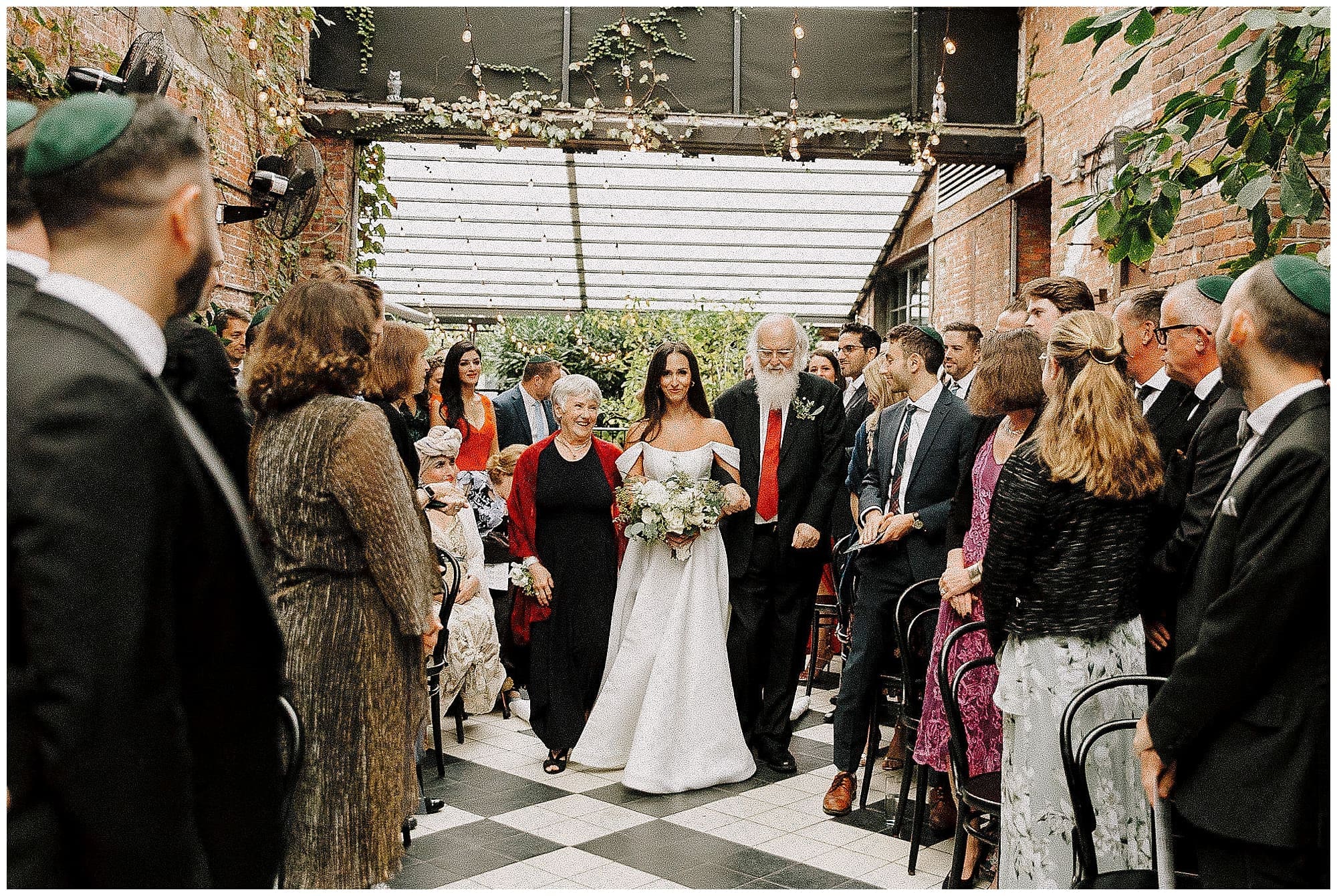 The bride walks down a crowded outdoor aisle during the processional at the Wythe Hotel in Brooklyn, New York. String lights and lush wedding florals overhead frame the ceremony space as guests look on.