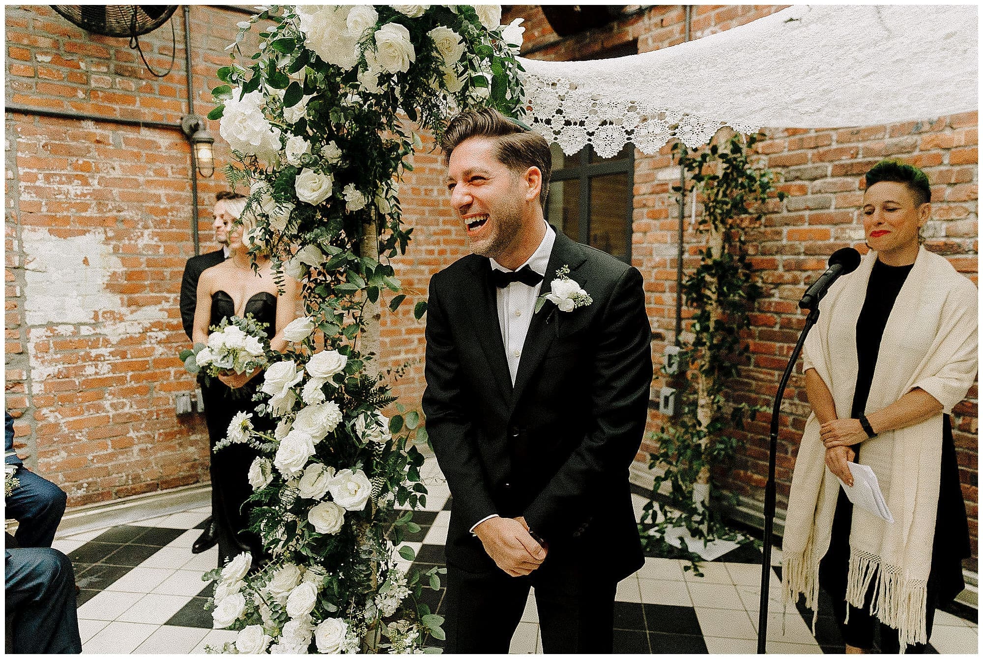 The groom stands at the altar laughing during his NYC wedding ceremony at the Wythe Hotel in Brooklyn, with an officiant wearing a white tallit beside him. A lush floral arch by a New York wedding florist and exposed brick wall are visible in the background.