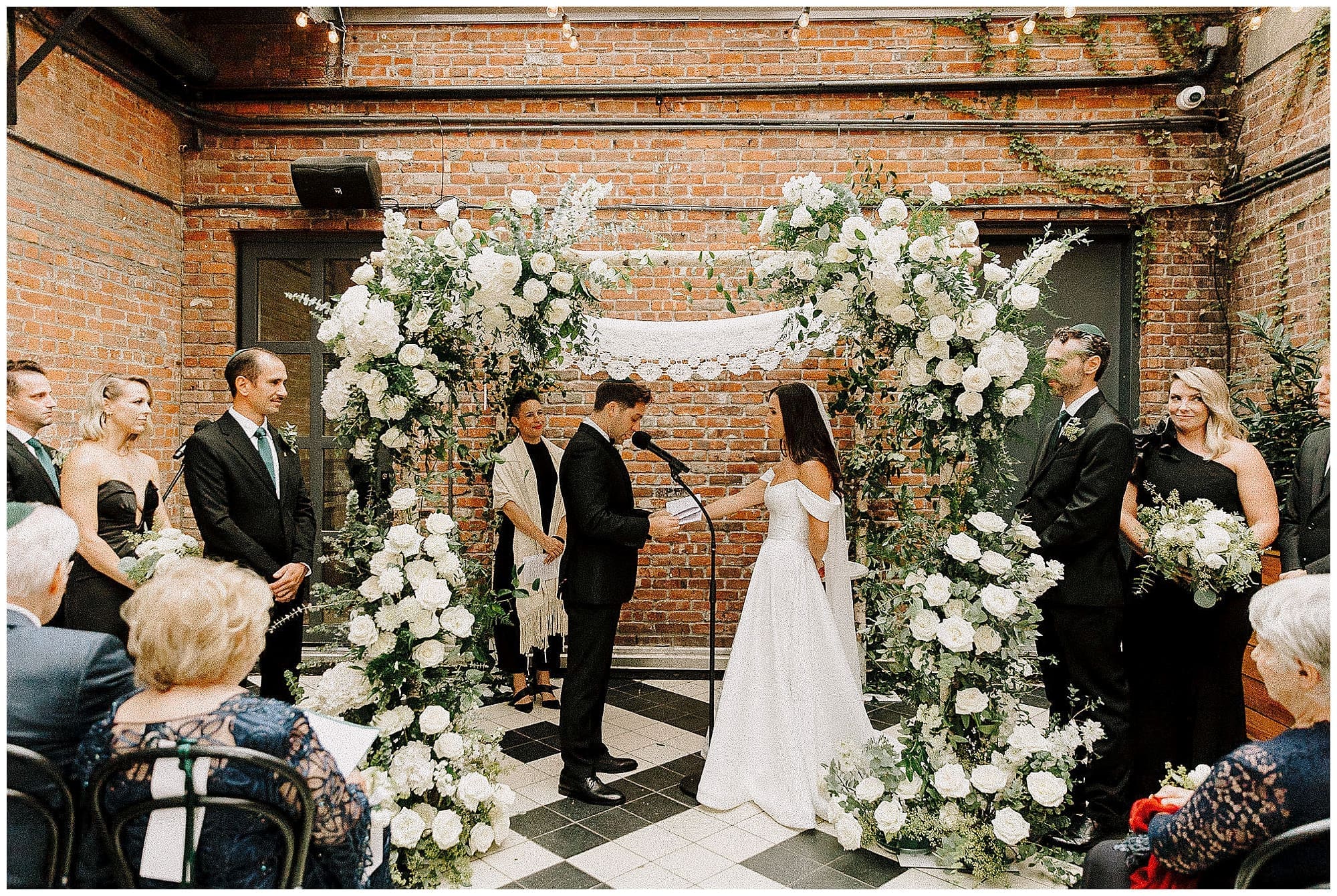 A wide shot of the ceremony space at the Wythe Hotel, Brooklyn, with guests seated in rows. The couple stands under a large wedding floral arch of white flowers and greenery crafted by a NYC wedding florist, set against an exposed brick wall.